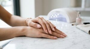 ידיים נשיות מטופחות עם מניקור לק ג'ל בצבע ורוד ניוד על שולחן שיש, ברקע מנורת ייבוש לק UV